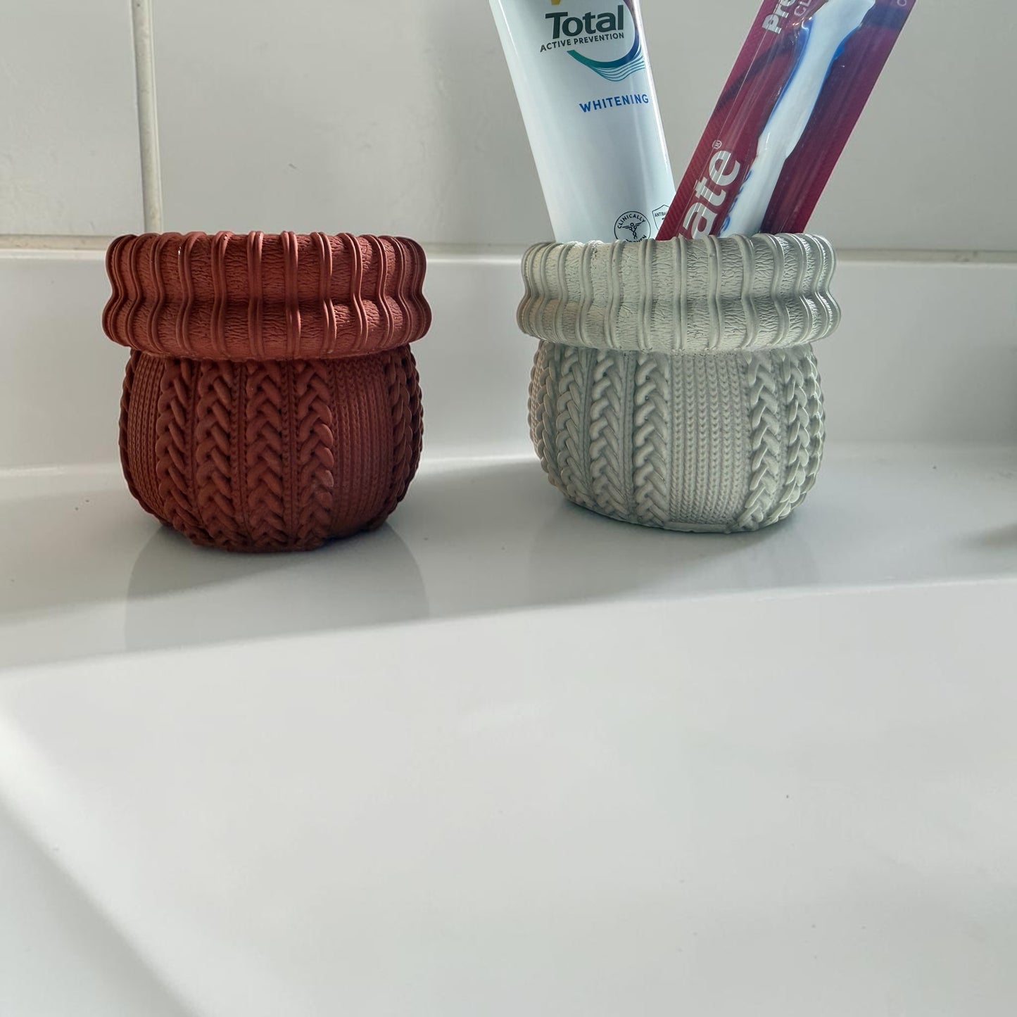 Two knitted containers holding toothpaste and toothbrushes on a white surface.